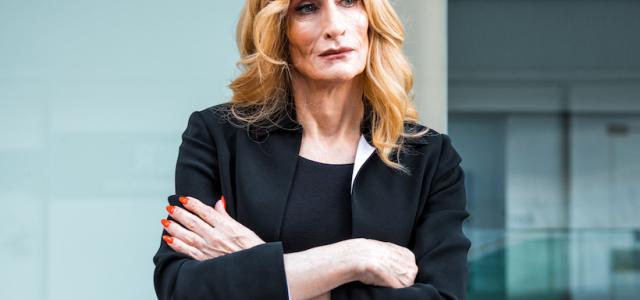 Transgender businesswoman with blond hair, a black suit, and a serious, pensive look on her face