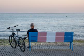 Older man with a bike next to him sitting on a beachside bench that is painted with the transgender pride flag colors
