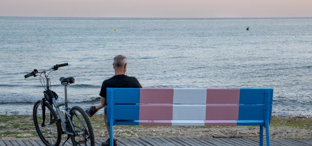 Older man with a bike next to him sitting on a beachside bench that is painted with the transgender pride flag colors